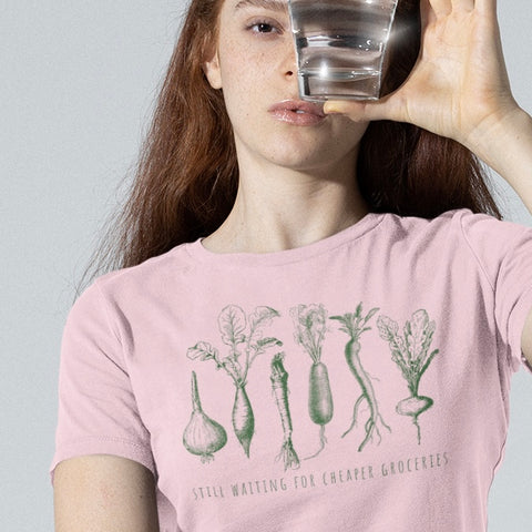 political t-shit that says still waiting for cheaper groceries in thin green writing featuring green illustrations of an array of root vegetables- shop feminist trash store for political clothing, feminist tees and acab t-shirts