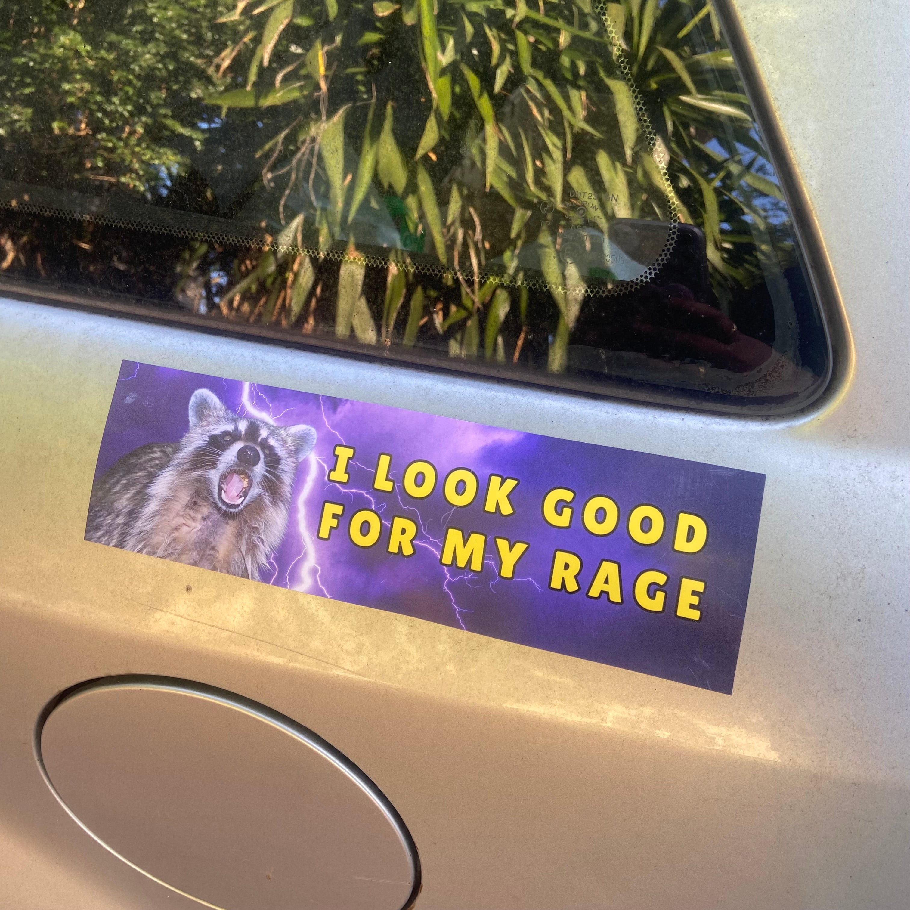 A close up view of a rectangular 10x3 inch car magnet featuring a roaring feral raccoon surrounded by bright purple lightning bolts with bold yellow text that reads i look good for my rage by Feminist Trash.
