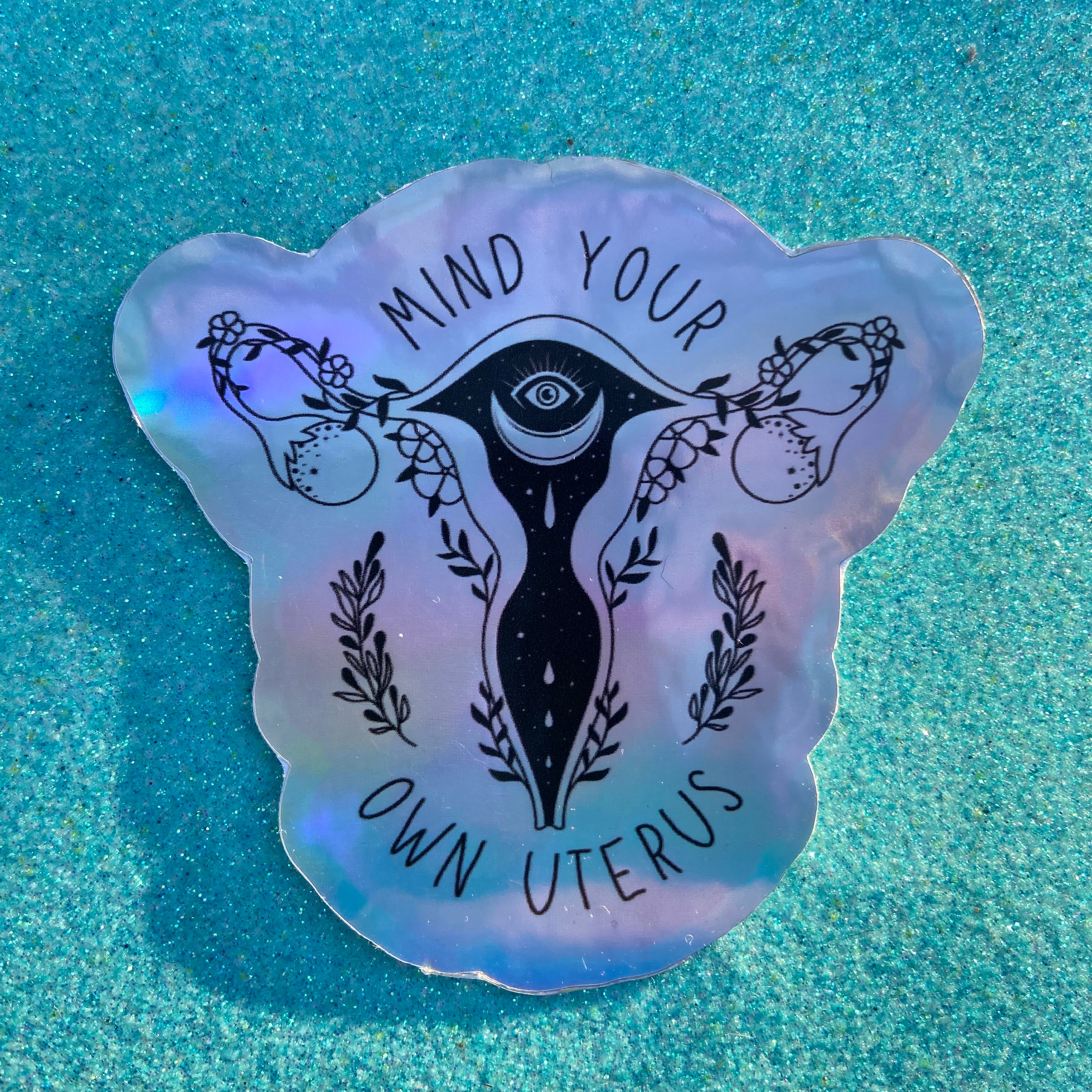 holographic feminist sticker that says mind your own uterus featuring a witchy uterus illustration- shop feminist trash for feminist stickers and political pins