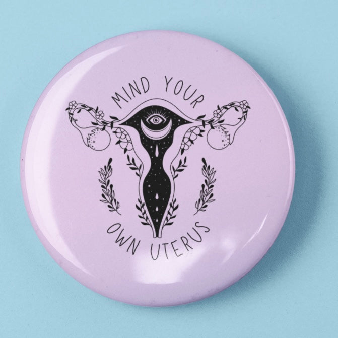 feminist pin that says mind your own uterus in black featuring a witchy uterus illustration- shop feminist trash for feminist pins and political tshirts