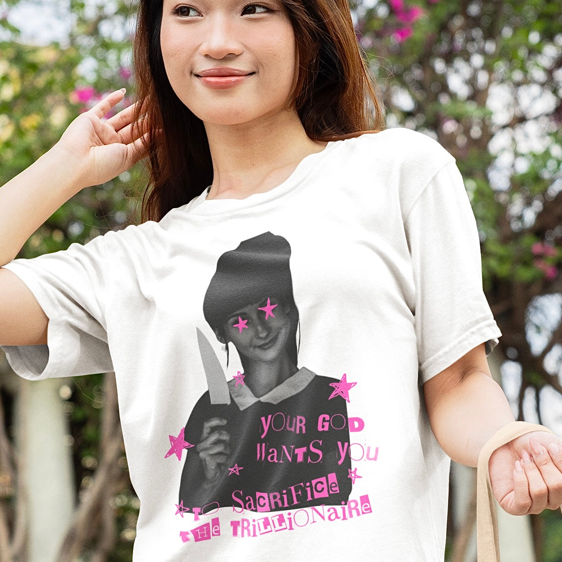 A smiling young woman wearing a white graphic political t-shirt by Feminist Trash that features a subversive black and white riot grrrl zine design reading "Your God Wants You to Sacrifice the Trillionaire" with hot pink stars over the graphic's eyes.