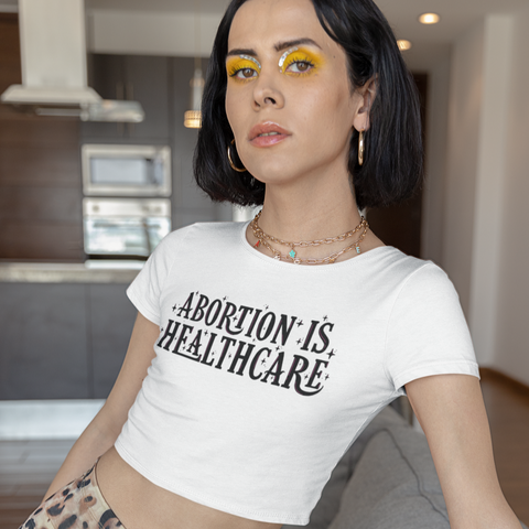 White feminist crop top that says abortion is healthcare in bold black writing with pink accents. - Shop social justice apparel and feminist tees