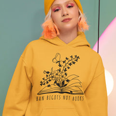 Gold/Bright Yellow Political hoodie that says ban bigots not books in black writing featuring an illustration of a book with flowers and a butterfly blooming out of the pages- shop feminist trash for political clothing, pride merch and feminist shirts. 
