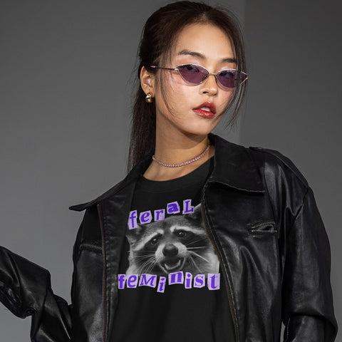 black raccoon tshirt that says feral feminist in purple ransom style writing featuring a black and white raccoon photography- Shop feminist trash for funny t-shirts for women and feminist clothing