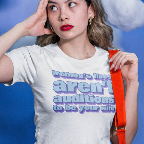 Image of a white feminist t-shirt with the phrase 'Women’s Lives Aren’t Auditions To Be Your Wife' in bold light blue text. This feminist tee is a powerful statement piece for anyone looking for feminist t-shirts, feminist tees, and feminist graphic tees. It embodies the spirit of feminism, making it an ideal feminist tshirt for those looking to express their beliefs boldly.