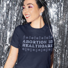 navy blue feminist tee that say abortion is healthcare in a festive snow christmas design- shop feminist trash for feminist tees and political clothing