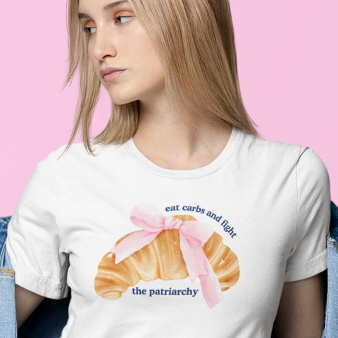 white feminist shirt that says eat carbs and fight the patriarchy in blue writing- featuring a coquette croissant watercolour illustration. Shop feminist trash store for feminist tshirts and dump him shirts