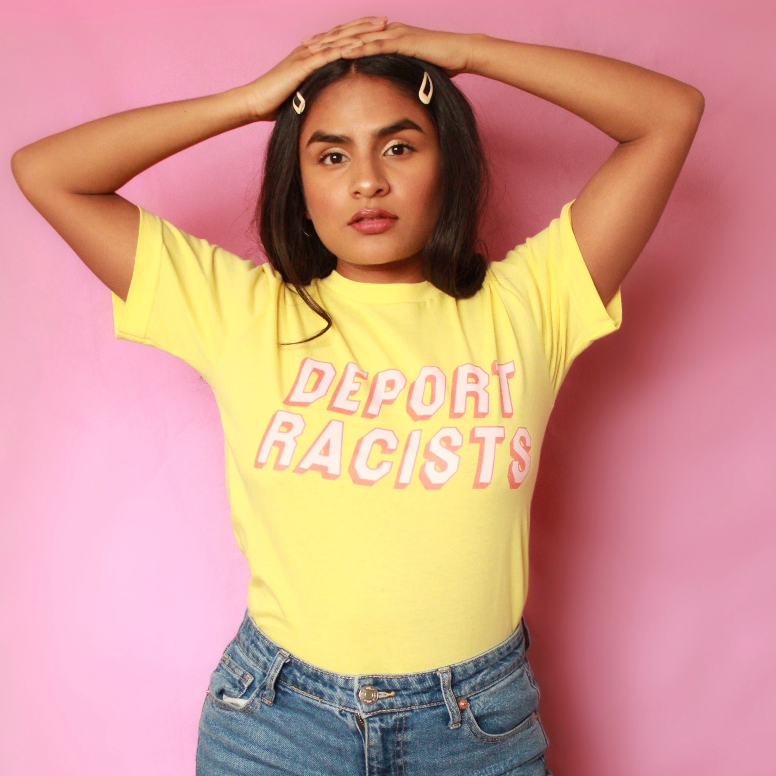 Empowering yellow feminist shirt featuring the message "Deport Racists" in peach writing