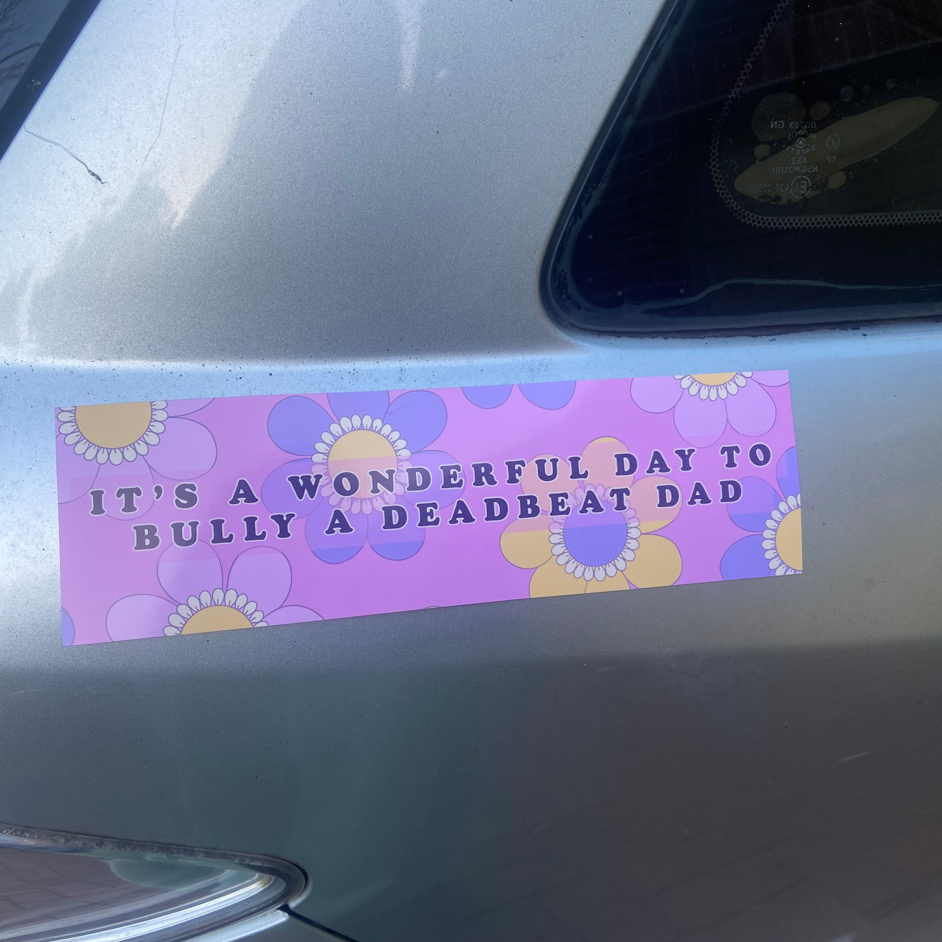 A close up view of a rectangular 10x3 inch car magnet featuring a cute retro light purple and yellow floral background with bold purple text that reads its a wonderful day to bully a deadbeat dad by Feminist Trash.