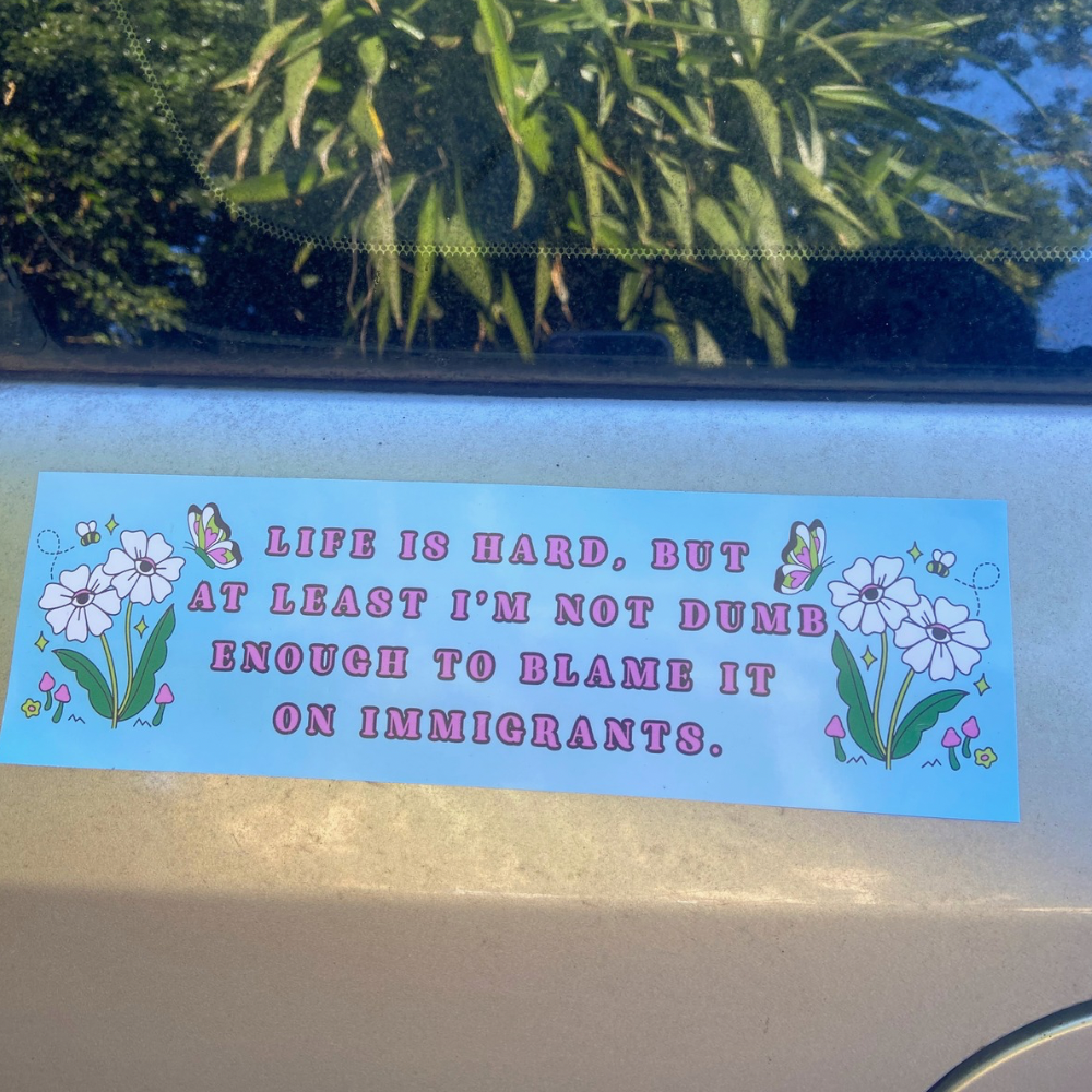 Life is hard but at least I'm not dumb enough to blame it on immigrants political car magnet with light blue background, white daisies, and butterflies
