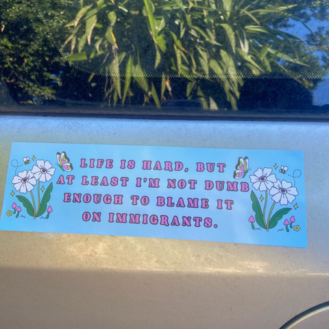 Life is hard but at least I'm not dumb enough to blame it on immigrants political car magnet with light blue background, white daisies, and butterflies