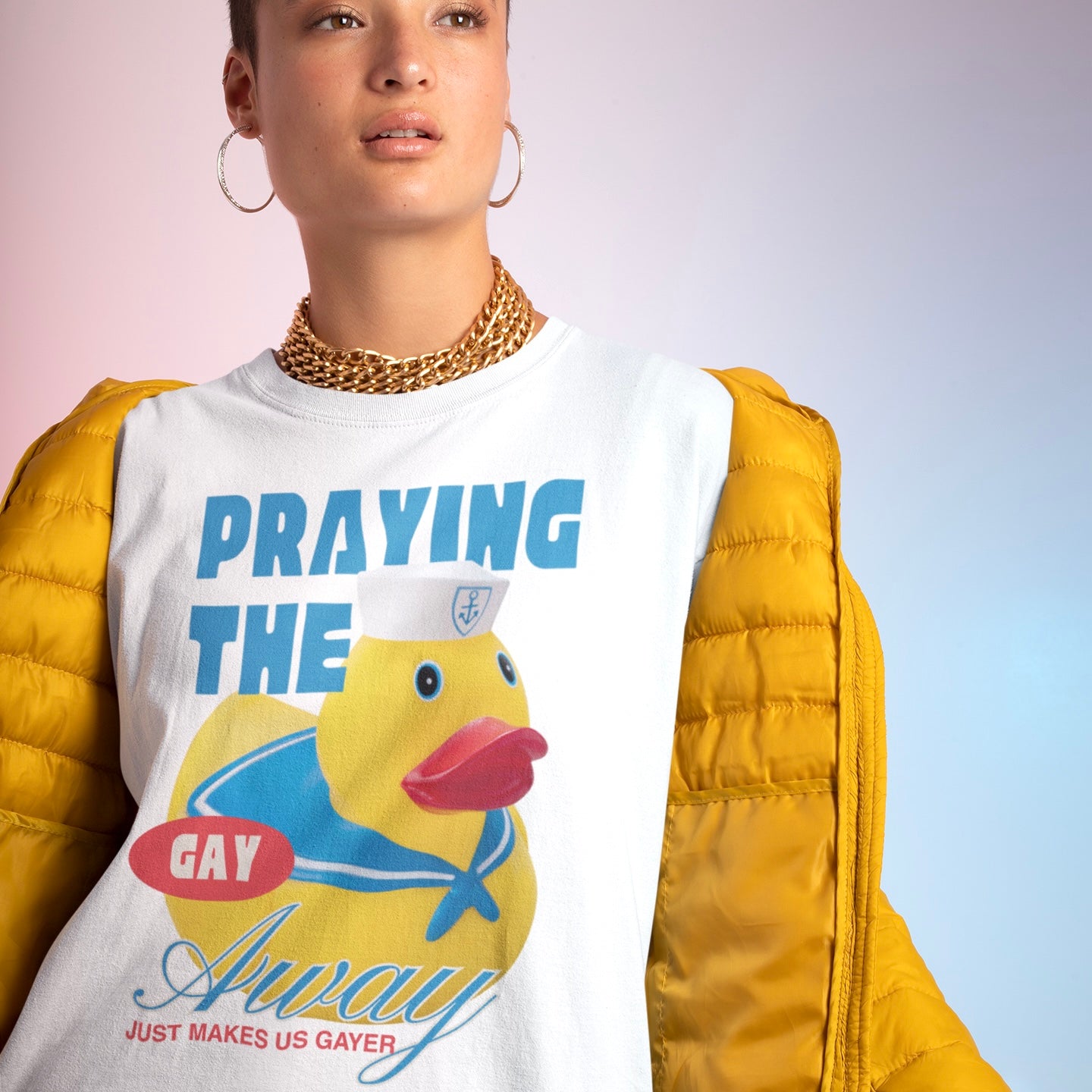 A person wearing the white Feminist Trash queer pride shirt that says praying the gay away just makes us gayer styled underneath a bright yellow puffer jacket with a gold chain necklace