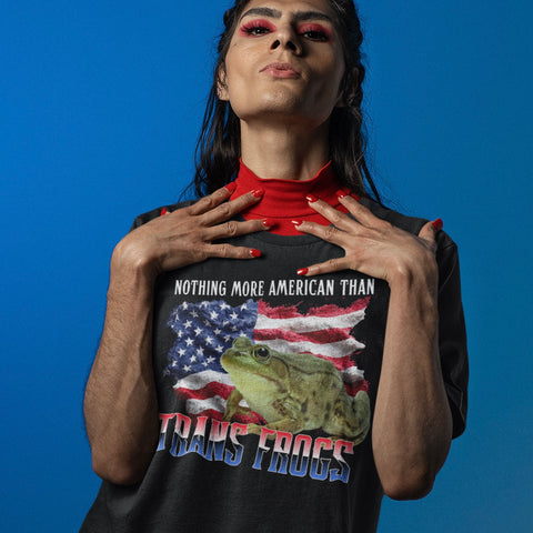 Close up look at a person modeling the black Trans Frogs Shirt From Feminist Trash,  layered over a bright red mock neck turtleneck. The individual has bold red eye makeup and matching red nails against a solid blue background. 